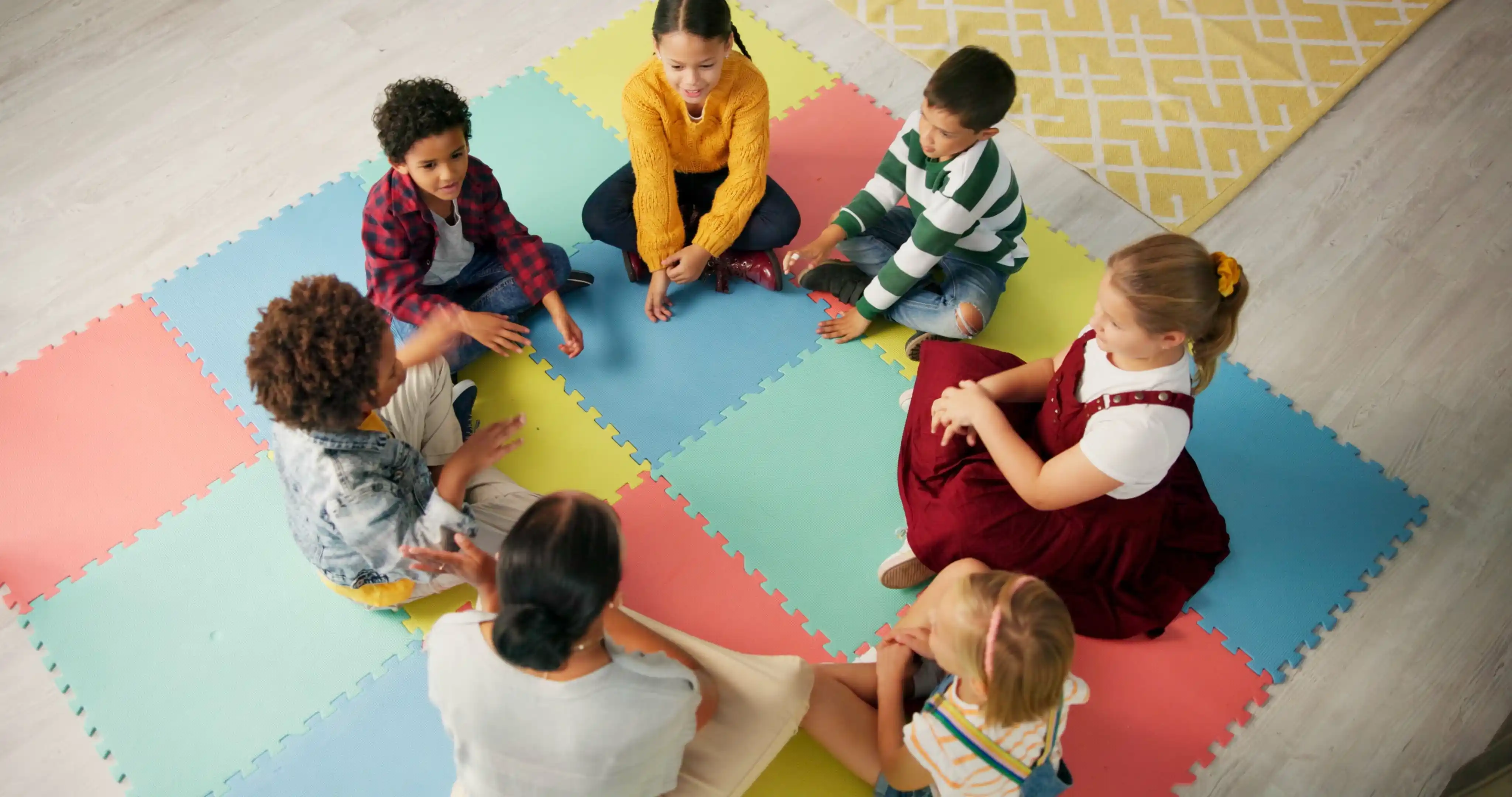 A group of children sitting in a circle on colorful foam mats, engaging in a group activity with a therapist.