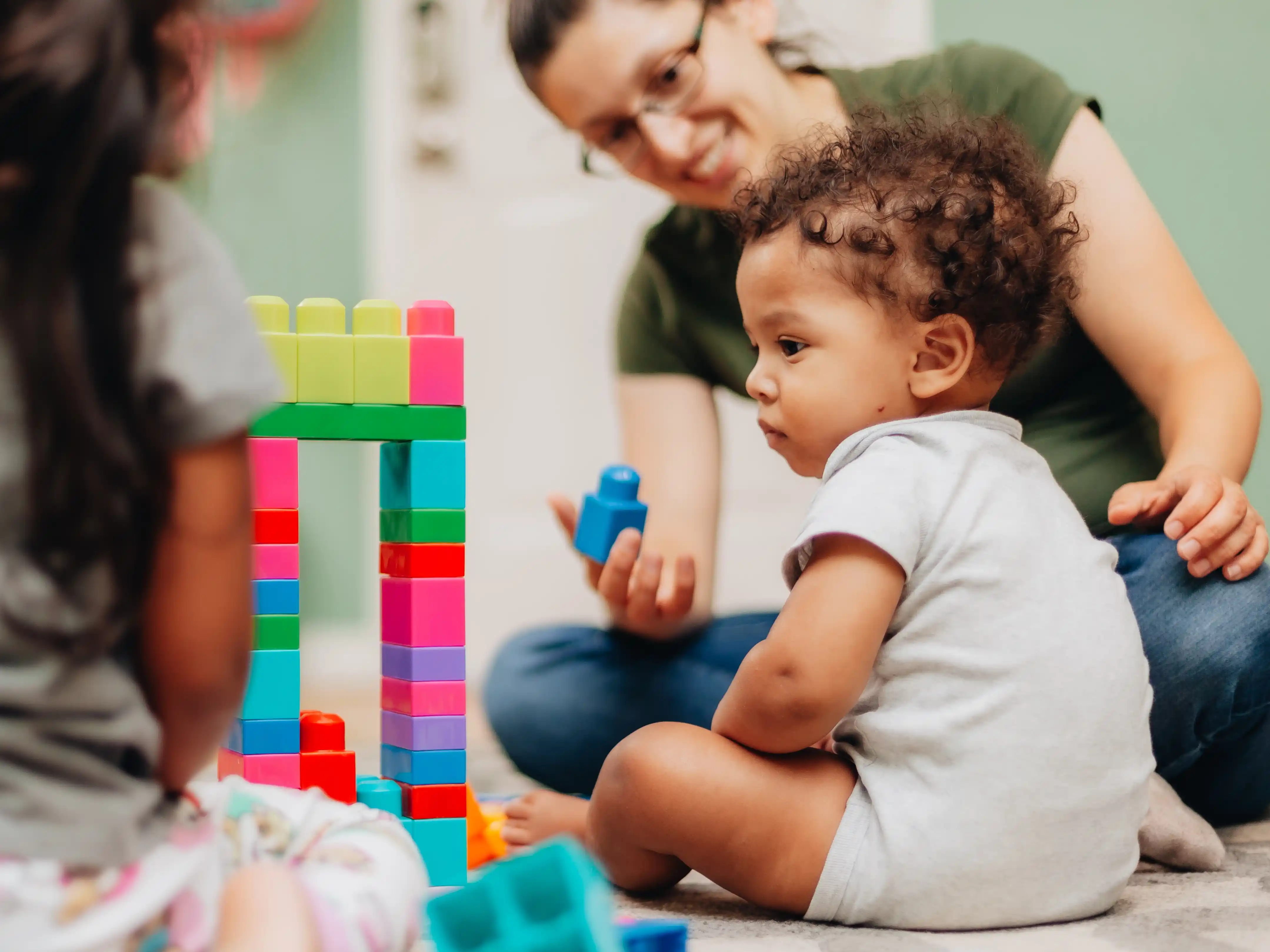 A toddler playing with colorful building blocks on the floor while a therapist watches nearby.